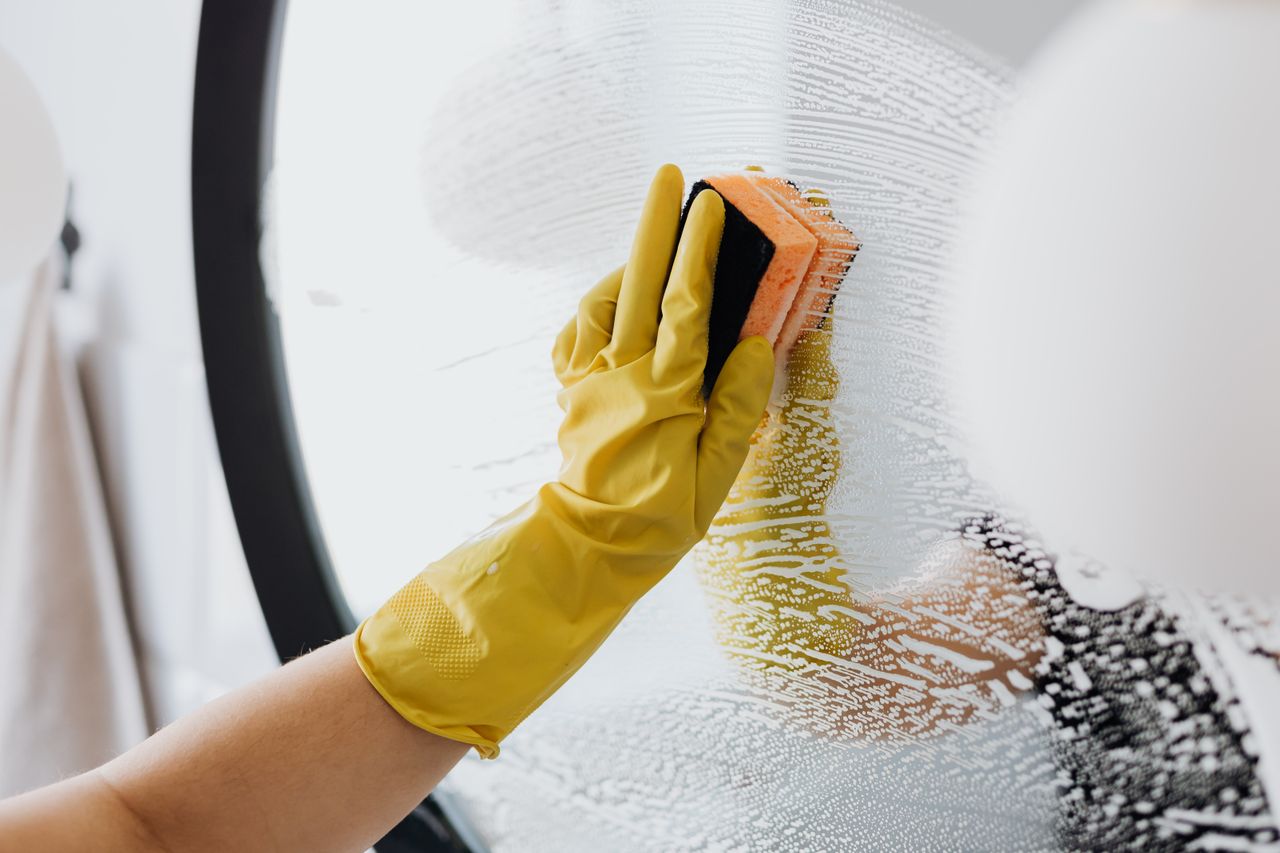 Personne anonyme portant des gants en caoutchouc jaunes et utilisant une simple éponge pour nettoyer un miroir dans une salle de bain